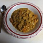 White bowl with a red stripe full of redish-brown creamy korma with chinks of chicken and tomatoes