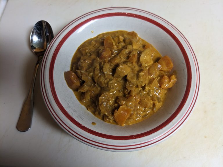 White bowl with a red stripe full of redish-brown creamy korma with chinks of chicken and tomatoes