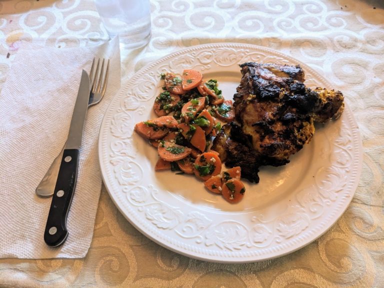 White plate with slightly charred chicken thigh. To the left is a small amount of carrot salad.