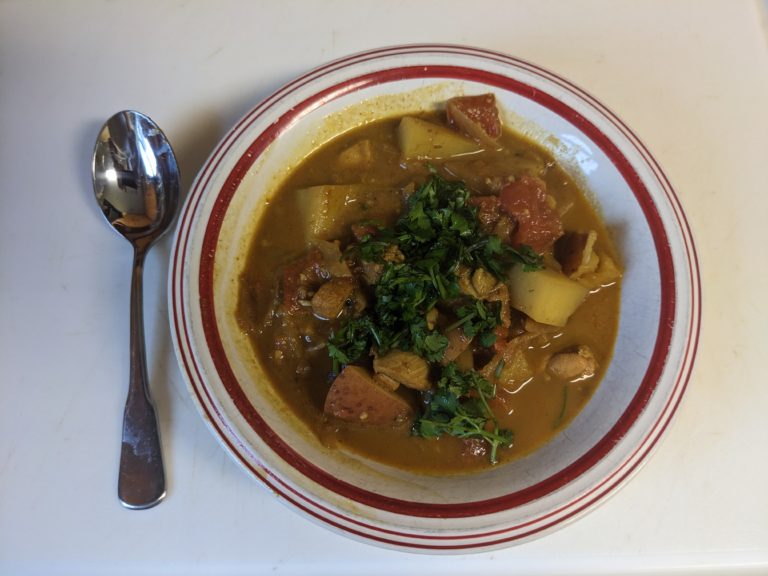 On a white background is a bowl with a red stripe around the rim. Inside the bowl are chicken and potato pieces in a reddish gravy and topped with a small pile of cilantro