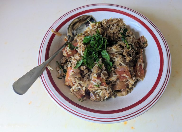 In a white bowl with a red ring around the rim is a combination of rice, meat, and tomatoes with a little cilantro leaves on top